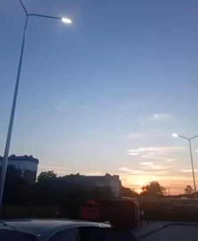 The video captures a daytime scene with a clear blue sky, a few wispy clouds, and the sun setting on the horizon. Two tall lampposts are visible, each with a light on. In the foreground, a portion of a car is visible, with its dark body and a hint of its interior. The background features buildings, trees, and what appears to be a parking lot with a paved surface. The overall atmosphere is serene and peaceful, suggesting an evening setting.