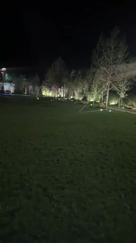 The video shows a nighttime scene outside a building with lights illuminating the exterior and trees. There is a large lawn in the foreground. Several spotlights are visible, some pointed at the trees and some at the building. Beams of light are also seen projecting from the building. The overall impression is of an event or gathering taking place.