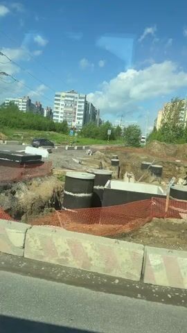 A video shows a roadside construction site. The site is bordered by orange safety netting and concrete barriers. A large excavation reveals concrete manhole sections and pipes. An orange Hitachi excavator sits on a mound of dirt within the site. In the background, apartment buildings rise under a blue sky with scattered clouds. Traffic passes on the road beside the construction.