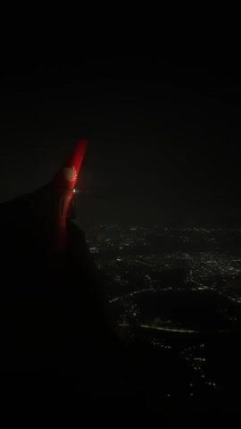 This is a point-of-view video taken from an airplane window at night. The camera is pointed downwards and towards the wing of the aircraft. The wing is dark red with a logo visible on its tip, which appears to be a lion's head. The airplane is flying over a city, which is illuminated by countless lights. The lights are spread out in a vast network, indicating a large urban area. The overall scene is dark, with the city lights providing the only significant illumination below. The flight appears to be steady, with no noticeable turbulence.