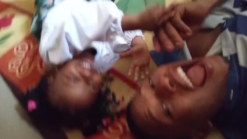 A man and a young girl are lying on a colorful patterned floor. The man is wearing a blue and white striped shirt, while the girl is wearing a white long-sleeved shirt. Both are laughing loudly. The girl has colorful beads in her hair. The camera is positioned above them, looking down.
