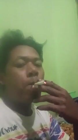 The video shows a close-up of a person's face as they smoke a cigarette. The person's face is the primary focus, with their mouth around the cigarette and smoke visible. Their hand holds the cigarette to their mouth. The background is a plain green wall. The person is wearing a white t-shirt with a colorful pattern. The lighting is dim, and the video appears to be taken in a low-light environment.