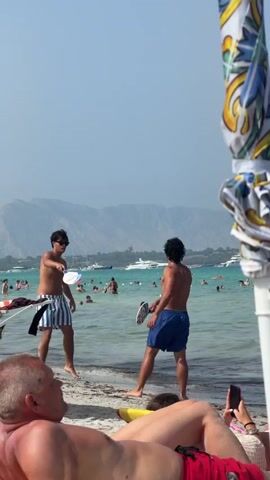 Two men are playing beach paddleball in the shallow ocean water. The man on the left is wearing blue and white striped shorts and the man on the right is wearing blue shorts. They are facing each other and holding paddleball rackets. Other people are swimming and relaxing on the beach in the background. The sky is clear and blue and there are mountains in the distance. A man in the foreground is lying on the beach, partially obscuring the view.
