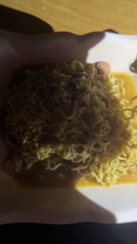 A plate of instant noodles with hot dogs and onions is shown. The noodles are tangled and covered in a yellowish broth. A fork is seen lifting a portion of the noodles. The plate is on a wooden table. The lighting is dim, casting shadows on the food.