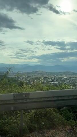 The video shows a view from a moving vehicle, likely a car, overlooking a landscape. The foreground features a metal guardrail, with trees and vegetation growing on the side of the road. Beyond the trees, a cityscape or town is visible in the distance, appearing somewhat hazy.  Further back, mountains are present, adding depth to the vista. The sky above is partly cloudy, with the sun shining brightly through the clouds, creating a diffused light. The overall impression is of a scenic view captured from a moving vehicle.