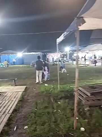 At night, a video captures a fairground or carnival scene. The camera moves around the event, showing various stalls and attractions. The scene includes food stalls with people working inside, rides adorned with colorful lights, and inflatable play areas. There are people, including children, wandering around. The ground is grassy, and the atmosphere is festive, lit by overhead lights.