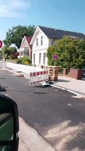 The video shows a street with ongoing construction. A car is moving forward, and the camera is mounted inside, capturing the view through the windshield and side mirror. The road is partially paved with asphalt, and there are construction barriers made of white and red plastic fences. Several traffic cones and hazard markers are also visible along the road. A speed limit sign indicating 10 is prominently displayed. Houses with traditional architecture, featuring red-tiled roofs and balconies, line the street. Trees and greenery are present, indicating a residential area. The sky is blue with some clouds, suggesting fair weather. The car is navigating through the construction zone, indicating a temporary traffic disruption. The overall scene depicts a street undergoing repair or renovation, with traffic being managed to ensure safety.