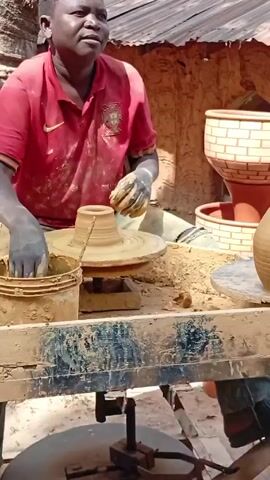 A potter in a red shirt sits at a pottery wheel shaping clay. The potter uses their hands and water to shape the clay into a pot. There is a bucket of clay next to the wheel. Finished pots are visible in the background. The potter is working in a covered outdoor space, possibly a workshop. The environment appears rustic, with a mud wall in the background.