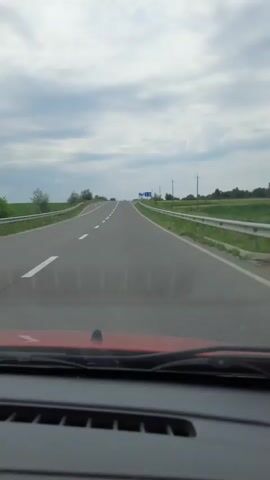 The video shows a view from the inside of a red car driving on a highway. The car is moving forward on a paved road with two lanes in the direction of travel and a shoulder on the right. The road is flanked by green grass and guardrails. There are signs visible on the right side of the road. The sky is overcast with clouds. The perspective suggests the video was recorded by a passenger or through a dashboard camera.