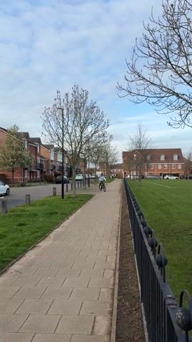 A person rides a bicycle along a paved path flanked by a grassy area on one side and a street with parked cars and buildings on the other. The path is bordered by a black metal fence on the grassy side. The person is wearing a light blue shirt and a hat. The sky is partly cloudy, and the trees are mostly bare, suggesting it is either late autumn or early spring.