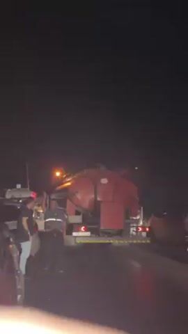 Here's a concise description of the video:

The video, shot at night, shows a roadside scene with several people standing near vehicles. At the center of the frame is a large, red truck with a tank-like container on the back. Additional vehicles, including a smaller truck and cars, are also present, suggesting a possible accident or roadside assistance situation. The road appears wet, possibly from recent rain. Several people are visible, and some appear to be wearing vests, possibly indicating they are roadside workers or emergency personnel. One of the people in the foreground is gesturing.