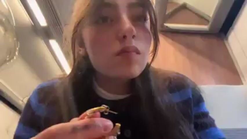 A young woman with light skin and brown hair tied loosely behind her head is sitting on a train. She is wearing a blue and black sweater over a white shirt and is putting on a small gold hoop earring. The train car has fluorescent lights and the interior is pale. The woman looks focused on her task.