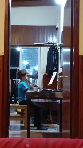 A young boy sits at a desk playing a wind instrument. He is wearing a blue t-shirt and black pants. The desk has sheet music on a stand and a laptop. The boy is looking at the sheet music. The scene is indoors, with wooden walls and a window in the background. There is a coat hanging on the wall behind the desk. A red fabric is visible in the foreground, partially obscuring the view.