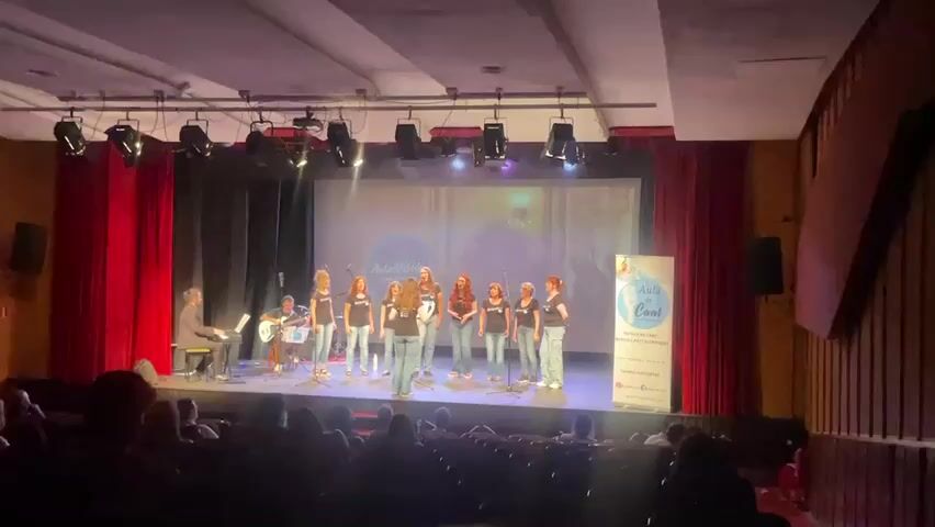 A group of nine young women, dressed in black t-shirts and blue jeans, are singing on a stage. They stand in two rows, with the front row slightly in front of the back row. Most of them are holding microphones. There is a man in a suit sitting at a keyboard on the left side of the stage, and another man behind him playing a guitar. A large screen is behind the singers, displaying a logo that says "Aula de Canto". To the right of the singers is a tall banner with text and logos. The stage is lit with spotlights, and red curtains hang on either side. An audience is visible in the seats in front of the stage, though their faces are not clear.