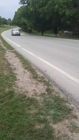 A silver sedan drives on a paved road lined with green grass and trees. The car is traveling from left to right in the frame. The camera appears to be positioned on the side of the road, slightly angled upwards. The sky is overcast, and the lighting suggests it is daytime.