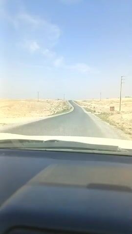 A dashboard camera perspective shows a winding asphalt road through a barren, desert-like landscape under a clear blue sky with sparse clouds. The road curves to the left and then to the right, disappearing over a distant hill. Power lines and poles are visible along the roadside, with low, scrubby vegetation sparsely scattered across the sandy terrain. The sun appears to be high in the sky, casting shadows and creating a bright, high-contrast scene. The vehicle's windshield and dashboard are visible in the foreground, indicating the camera is inside a moving vehicle. There are no other vehicles or people visible in the frame.