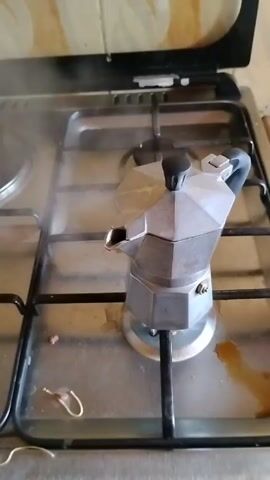 An eye-level, close-up shot shows a Moka pot on a gas stove. The Moka pot is silver with a black knob on top of its lid and a black handle on its side. It sits on the black grates of the stove, which is made of stainless steel. Steam is rising from the pot, indicating that it is brewing coffee. Next to the pot, there is a small spot of spilled coffee and a gold-colored paperclip. An electric stovetop burner is visible to the left of the Moka pot. The backsplash behind the stove has a patterned tile design.