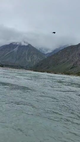 This video shows a wide, slow-moving river in the foreground. The water is a murky gray-blue and appears calm with slight ripples. In the background, a vast mountain range rises dramatically. The mountains are rugged and steep, with patches of green vegetation clinging to their lower slopes. The upper portions of the mountains are shrouded in thick, white clouds, obscuring the peaks and creating a dramatic, overcast atmosphere. The lighting is dim, consistent with a cloudy or overcast day. The camera perspective is low, as if at water level, and it appears to be panning slowly from right to left, revealing more of the mountain landscape. There are no visible people or man-made structures in this segment of the video.