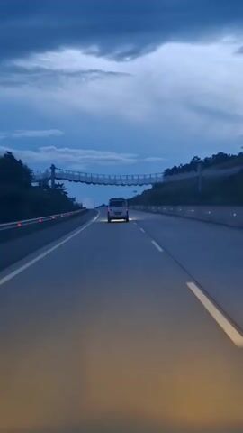 A van is driving down a highway at dusk. The sky is dark blue with some clouds. There is a suspension bridge over the highway. The road has yellow and white lines. The van's headlights are on.