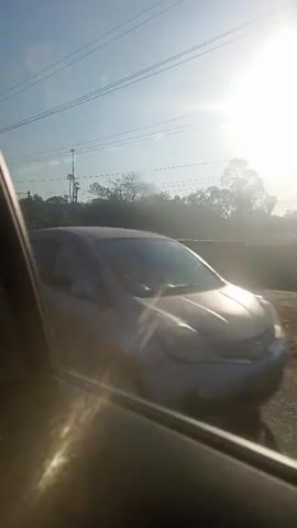 This video appears to be captured from inside a moving vehicle, looking out the side window. The sun is bright and positioned high in the sky, casting a strong glare and lens flare across the scene. The background shows a rural or semi-rural environment with trees and vegetation. There are utility poles and power lines visible against the bright sky. A dirt or gravel road runs parallel to the vehicle's path, and beyond that, there is more greenery and possibly some low-lying structures. Dust or light particles are visible in the air, suggesting a dry environment or the movement of the vehicle kicking up dust. In one frame, a white car is seen passing by in the opposite direction. A small white object, possibly a piece of paper or debris, is briefly visible in the sky. The overall impression is of a journey through a sunny, somewhat dusty landscape.