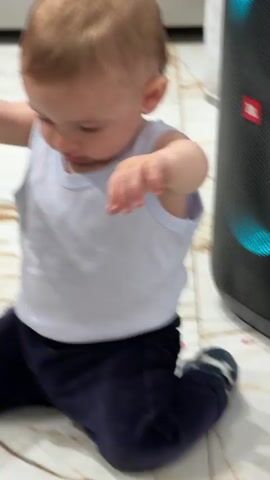 A baby is sitting on a tiled floor. He is wearing a white tank top and dark pants with a blue sock on one foot. A speaker is next to him. The baby turns his head. Another child walks into view, wearing a pink striped shirt and having their hair in pigtails. The child is looking with a slight grimace. The baby crawls behind her.