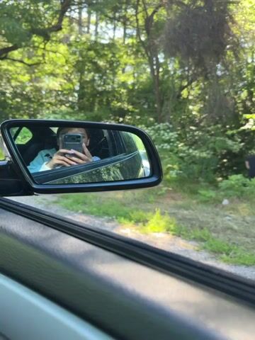 A video shows a man urinating in the woods from the perspective of someone in a car. The video is composed of three clips. The first clip is a wide shot of the woods with a man urinating in the background. The second clip zooms in on the man. The third clip shows a reflection in a car mirror of a person taking a selfie. The video has a total duration of 5 seconds.