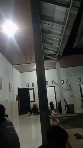 The video shows an outdoor area with several people sitting and interacting. The environment appears to be a courtyard or patio with a roofed structure overhead, possibly part of a building. The lighting suggests it is either evening or nighttime. Several individuals are visible, some sitting on the ground, while others are closer to the building. Objects in the scene include doors, windows, and a trash can. The overall atmosphere seems casual and social, with people gathered together.