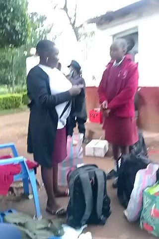 The video depicts an outdoor scene in what appears to be school grounds in Ngenyilel ward, Kenya. The setting is on a dirt surface with exposed tree roots.

Multiple individuals are present, primarily young women or girls, many dressed in maroon skirts or dresses, suggesting school uniforms. An adult woman, wearing a white top and a dark cardigan or jacket over a maroon skirt, is centrally featured. She is seen examining or rummaging through items, holding a black piece of clothing at the beginning of the video, and later bending down to inspect the contents of a black backpack.

Clustered around the individuals are various bags and belongings on the ground, including several black backpacks, colorful patterned tote bags, and striped plastic bags. Also visible are small white cardboard boxes and a piece of blue fabric. In the background, parts of a light-colored building and a red crate can be seen. Another person, partially obscured and wearing a face mask, is briefly visible behind the adult woman. The legs and feet of a person wearing light-colored shoes are visible in the foreground, suggesting someone is seated nearby.

The activity suggests an inspection, packing, or unpacking process, possibly related to school events, travel, or dorm arrangements. The individuals appear focused on their tasks. No clear, legible text is visible on any objects or in the background.