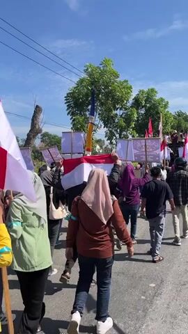 A group of people are marching outdoors on a paved road under a bright blue sky with some clouds. Many are holding Indonesian flags, which are red and white. Some individuals are carrying signs, one of which is a large pink poster with handwritten text in black. The text on the pink sign appears to say "KAMI MENDUKUNG PROGRAM MAKANAN BERGIZI GRATIS" which translates to "WE SUPPORT THE FREE NUTRITIOUS FOOD PROGRAM." Some people are wearing headscarves and casual clothing. Trees and buildings are visible in the background. The overall scene depicts a peaceful demonstration or parade.