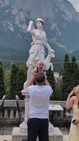 A man holds a baby up towards a white marble statue of a woman with a deer. A woman stands to the right, looking towards the man and baby and holding her hand near her mouth. The statue is on a pedestal in front of a lush green forested mountain range. Cypress trees are planted in a row behind the statue. A wrought iron gazebo structure is visible on the right side of the frame.