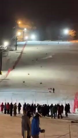 A crowd of people watches someone ski down a snow-covered slope at night.  There are lights illuminating the slope, and a ski lift is visible on the left side of the frame. The skier is making their way through a course marked with red and blue flags.  There are orange or red safety nets along the side of the slope. The sky is dark. The group of people watching the skier are standing on a lower section of snow, and many of them appear to be wearing winter clothing.