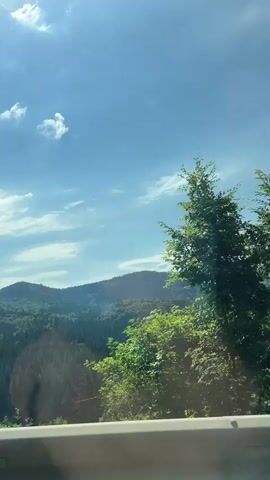The video captures a scenic view of lush green mountains and trees under a bright blue sky with scattered white clouds. The perspective suggests the footage is taken from a moving vehicle, possibly a car, as evidenced by the slight blur and the presence of what appears to be a windshield or window in the foreground. Sunlight filters through the trees, creating dappled light and occasional lens flares. The overall impression is one of a peaceful journey through a natural, verdant landscape.
