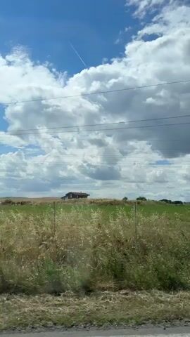 The video shows a view from a moving vehicle, likely a car, looking out over a rural landscape under a partly cloudy blue sky. In the foreground, tall dry grass and some green vegetation are visible. A fence line with wooden posts runs through the middle ground. In the distance, there are fields of golden grain, a few trees, and what appears to be a small, dilapidated structure with a visible roof and posts. Power lines stretch across the upper portion of the frame. A contrail from an aircraft is visible in the sky. The scene suggests a journey through the countryside on a bright, possibly windy day.