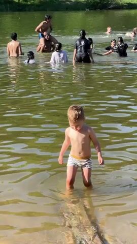 A young boy in striped swim trunks is waist-deep in a body of water and waving. Behind him, several other people are also in the water. Some are swimming while others stand. One man is sitting on another man's shoulders. The water is murky and greenish. Trees line the shore in the background. The day appears to be sunny.