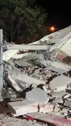 The video shows the aftermath of a building collapse at night. Debris, including concrete, bricks, and furniture, is scattered across the site. The building's structure is severely damaged, with walls and floors completely destroyed. A street light illuminates the scene, and trees are visible in the background.