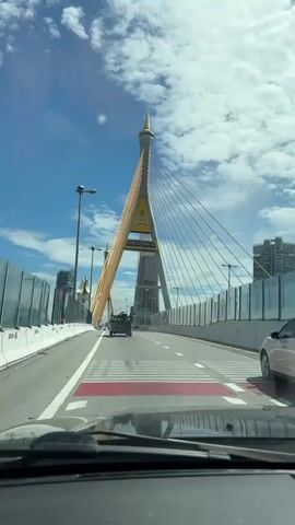 The video shows a view from inside a vehicle as it travels across a suspension bridge. The camera is positioned behind the windshield, allowing a clear view of the road ahead. The bridge's signature cable-stayed design is prominently visible, with thick yellow cables extending from a towering central pylon to the bridge deck. The pylon is triangular in shape and topped with a golden spire. Below the pylon, a sign with Thai writing and an emblem is displayed. On the right side of the road, a white car is driving in the same direction. On the left side of the road, a white barrier separates the lanes from the other side of the bridge, which is partially obscured by a fence. Several streetlights are visible along the bridge. In the distance, a tall, unfinished skyscraper is under construction, and other buildings can be seen. The sky is blue with scattered white clouds. A dark-colored truck is driving ahead of the vehicle. The overall impression is a daytime journey across a modern urban bridge.