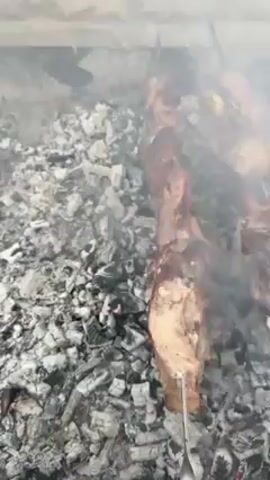 The video shows meat skewers cooking over hot coals in what appears to be an outdoor brick barbecue setup. The meat is arranged on metal skewers, and the camera pans across them, showing the different pieces as they cook. There is a lot of smoke rising from the coals, partially obscuring the view of the meat. The background shows a stone wall, indicating an outdoor setting.