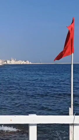 The video is an outdoor scene with a bright blue sky and the deep blue ocean. In the foreground, there is a white railing, and a red flag is attached to a white pole. The flag is blowing in the wind. In the distance, there is a coastline with buildings. The water is choppy.