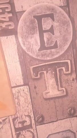 The video shows a close-up, angled view of what appears to be a piece of art or a textured surface. On the right side of the frame, there are metallic letters that spell out "ET" with rivets visible around the "T". To the left of the letters is a vertical strip with black numbers "345685" printed on it. The majority of the frame on the left displays a colorful painting of two figures on a camel in a desert landscape, with a stylized sun or moon in the sky and reddish-brown desert formations in the background. The painting has a textured appearance, possibly made with sand or a similar material. The lighting suggests it is an indoor setting.