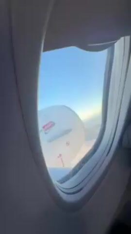 From the window of an airplane, the exterior is visible. The wing of the plane, white with a yellow and black striped portion and arrows, is partially visible. The sky is blue and there are some clouds.
