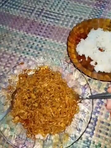 This is a close-up, top-down video of a meal being prepared or served. The main focus is on a plate of instant noodles, seasoned and mixed, with some visible bits of what appear to be egg or chicken. The noodles are a deep brown color, suggesting they are seasoned with a savory sauce. To the upper right of the frame, there is a smaller, ornate brown bowl filled with white rice. The meal is presented on a textured placemat with a woven pattern in various shades of purple, blue, and beige.

A fork is actively engaged with the noodles, suggesting someone is in the process of mixing them or serving them. In the latter part of the video, a hand with an orange fingernail is visible, holding the fork. The lighting appears to be artificial, suggesting the meal is being prepared or consumed indoors. There is no visible text in the video. The scene is simple and domestic, focused entirely on the food. The time of day and weather are not discernible.
