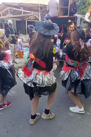 This is a vertical, eye-level video of a street performance. There are at least three people visible in the foreground, dressed in elaborate costumes. The costumes appear to be made of black fabric, possibly skirts, layered with shiny silver and red materials. They also wear pointed hats and have long, dark hair, giving them a witch-like appearance. One person in the front is wearing red sneakers, while another in the back is wearing white sneakers. Their actions suggest they are dancing or performing a choreographed routine. In the background, there are spectators and structures that appear to be shops or stalls, indicating a public event or festival. A large speaker system is also visible on the left side of the frame, suggesting music is accompanying the performance. The environment appears to be a street with paved ground and a bright, sunny day.