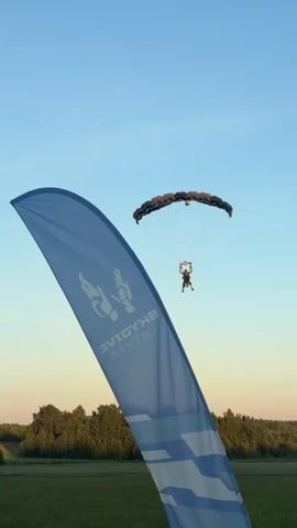 A tandem skydiver is captured descending towards a grassy field with a red airplane visible on the ground. In the foreground, a large blue flag with white graphics and text is prominently displayed, partially obscuring the skydiver and airplane. The sky is a clear blue with a hint of orange near the horizon, suggesting either sunrise or sunset. The skydiver is attached to a parachute and appears to be preparing for landing.