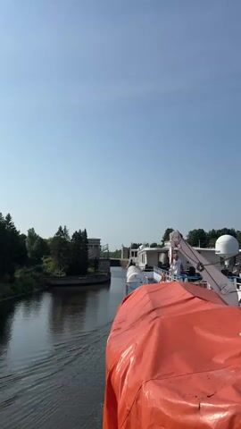 A river cruise ship is approaching a lock gate on a clear, sunny day. The ship is visible in the foreground, with an orange tarp covering part of its deck. The lock gate is a concrete structure with two towers, one on each side of the channel. Two statues are visible on top of the left tower. The water in the river is calm, and the sky is clear blue. The trees on the riverbank are green and lush.