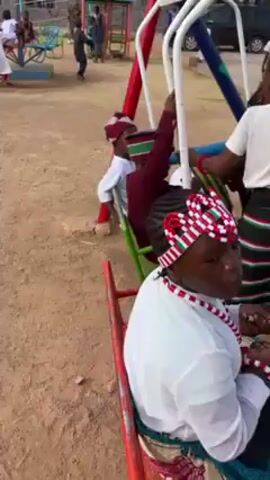 The video shows a group of children on a playground carousel. They are wearing traditional Nigerian clothing. The carousel is painted in bright colors, including red, white, green, and blue. Some children are seated, while others hold onto the railings. One child is facing the camera, smiling and laughing. The playground is sandy, and other people are visible in the background.