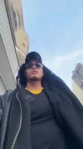 A young man wearing a black hooded jacket, a yellow t-shirt, a black cap with silver accents, and sunglasses walks down a city street, holding and smoking what appears to be a small electronic cigarette. He occasionally looks into the camera. There are parked cars on the street and buildings on either side of the sidewalk. The sky is visible and is a light blue.