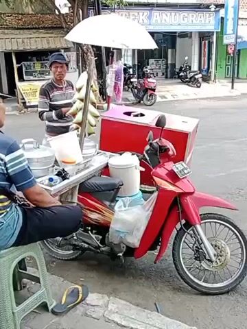 A man is standing next to a red motorcycle that is equipped with a mobile food cart. The cart has a pink cooler on the back of the motorcycle and a white umbrella providing shade. There are several metal containers on the cart, one with a white lid. A second man is sitting on a stool to the left of the cart, facing away from the camera. The scene appears to be on a street, with buildings visible in the background. One building has a sign that reads "OTEK ANUGRAH". There are other motorcycles and people in the background, suggesting a public area.
