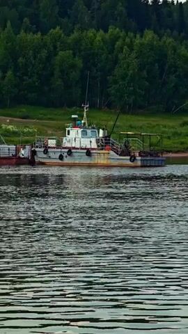 A small vessel is docked on a river. The vessel is gray and white with some rust. It has a cabin with windows and a mast. There is a separate barge attached to the front of the vessel. The river has choppy water. In the background is a dense forest of green trees. The sky is cloudy.