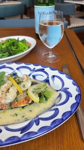 The video pans across a wooden table setting at a restaurant. In the foreground, a white plate with a blue and white patterned rim holds a dish of pan-seared fish topped with shrimp, lemon slices, and parsley, all covered in a creamy sauce. To the left, a white oval plate contains steamed green vegetables, likely broccolini, with a drizzle of sauce. Behind the food, a glass of water with ice sits next to a green bottle of S.Pellegrino sparkling natural mineral water. Further in the background, glimpses of restaurant seating with blue upholstery and wooden chairs are visible. The lighting is natural and appears to be daylight.