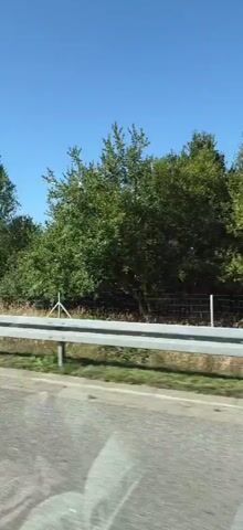 The video is a side-view shot of a wind turbine on a clear, sunny day. The turbine is white with red tips on its blades. It is situated in a green field with a few trees in the background. A road with a guardrail runs in the foreground, suggesting the video was filmed from a moving vehicle. The sky is blue with a few scattered clouds.